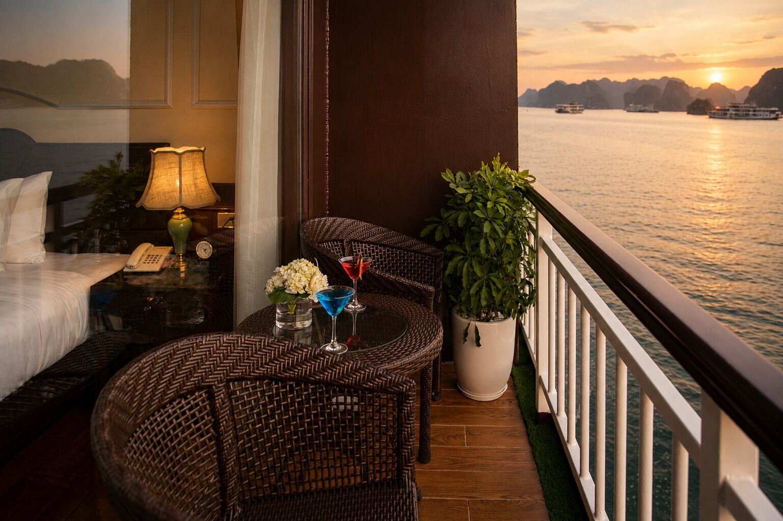 A balcony with wicker furniture, a potted plant, and a view of the water at sunset.