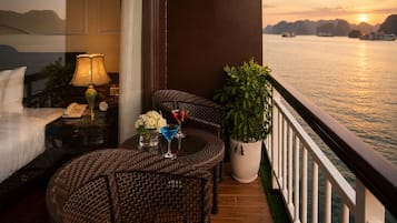 A balcony with wicker furniture, a potted plant, and a view of the water at sunset.
