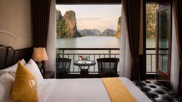 A hotel room with a bed, a nightstand, a lamp, a table with chairs, and a balcony with a view of a body of water and cliffs.