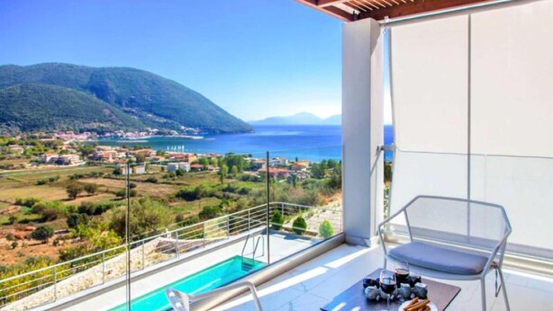 A balcony with a pool, a table with drinks, and a view of a coastal town. - Omicron & White Villas (Lefkada)