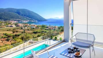 A balcony with a pool, a table with drinks, and a view of a coastal town.