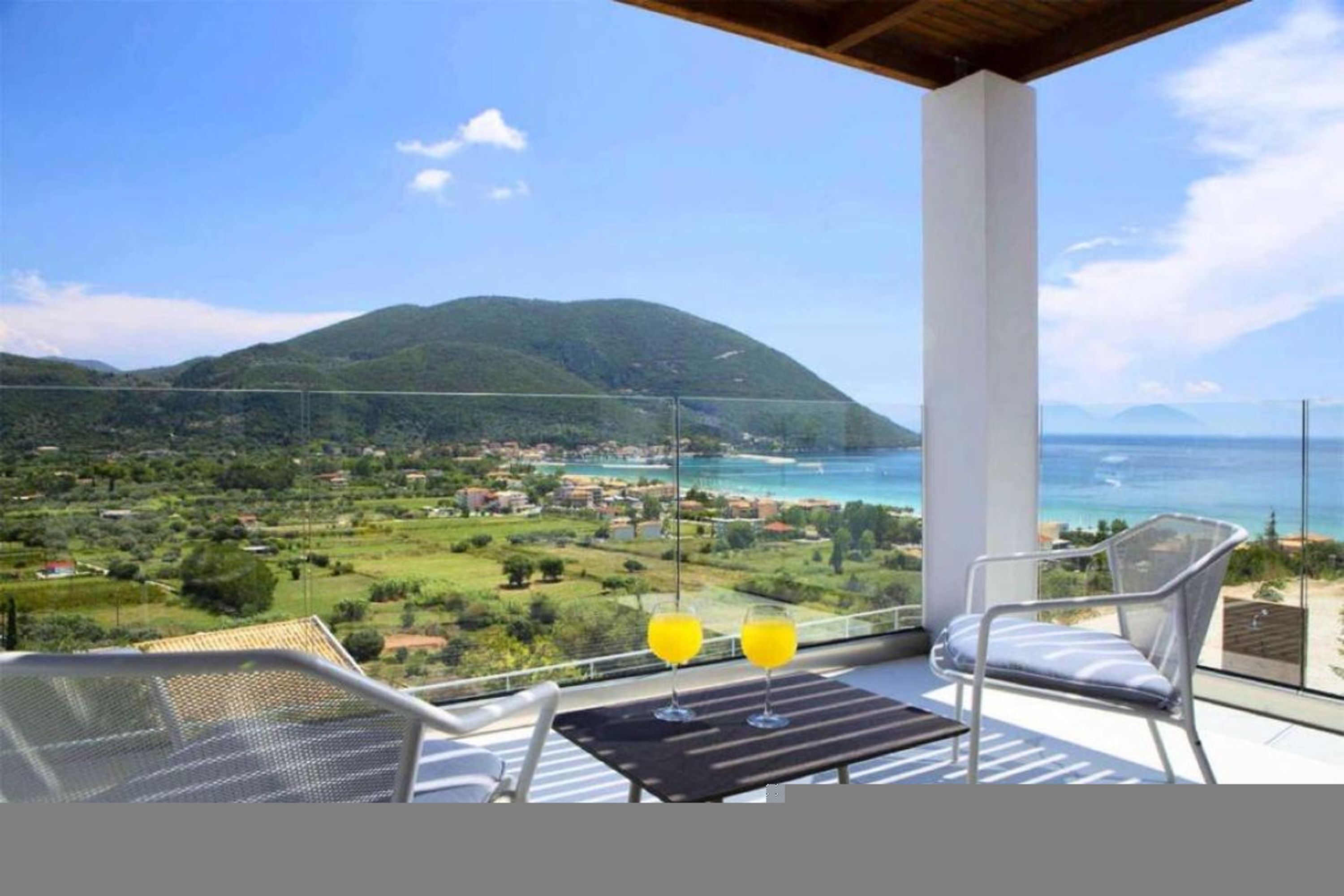 A balcony with a table and chairs, overlooking a coastal landscape with mountains and a town.