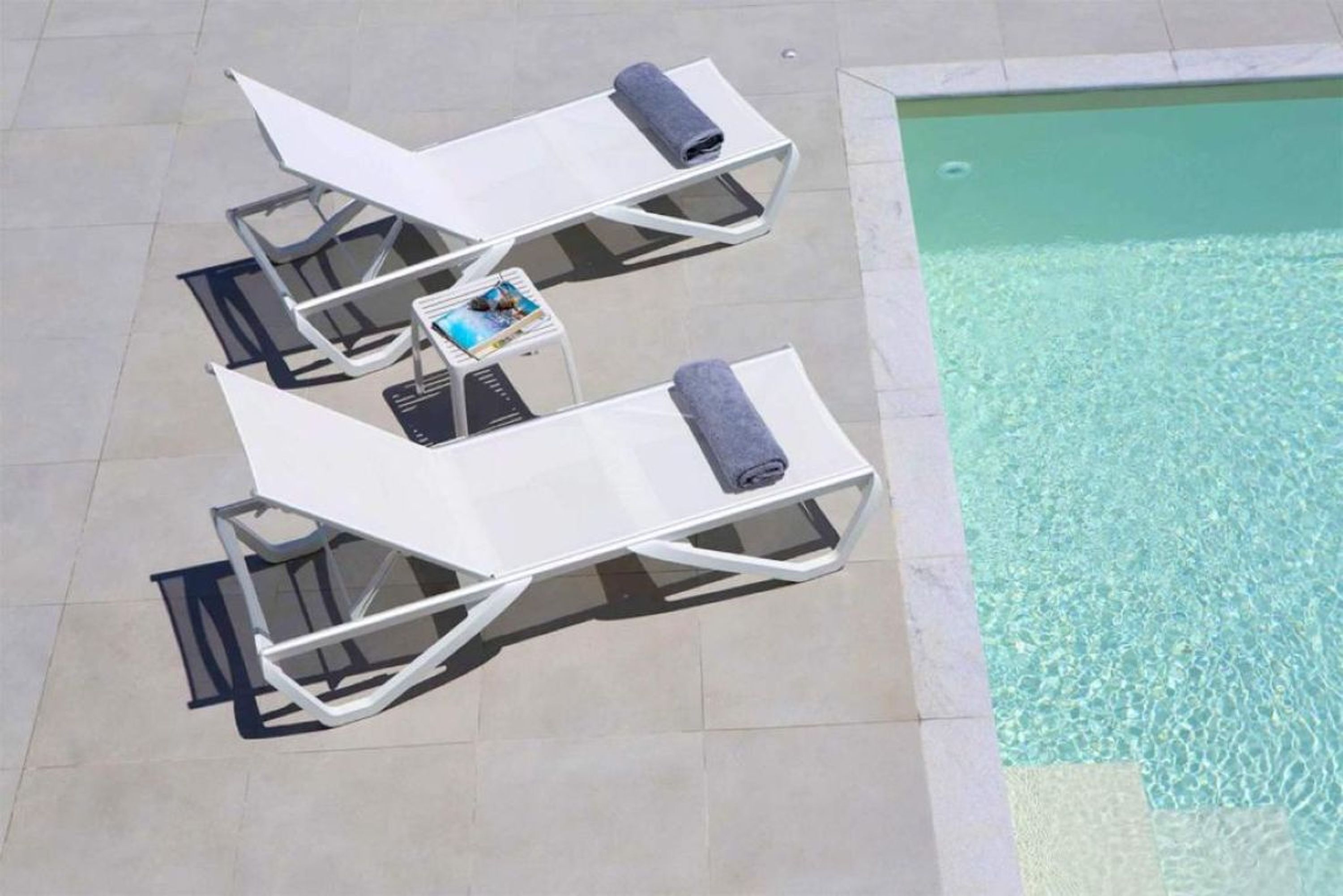 A poolside area with two white lounge chairs and a small table with a magazine.