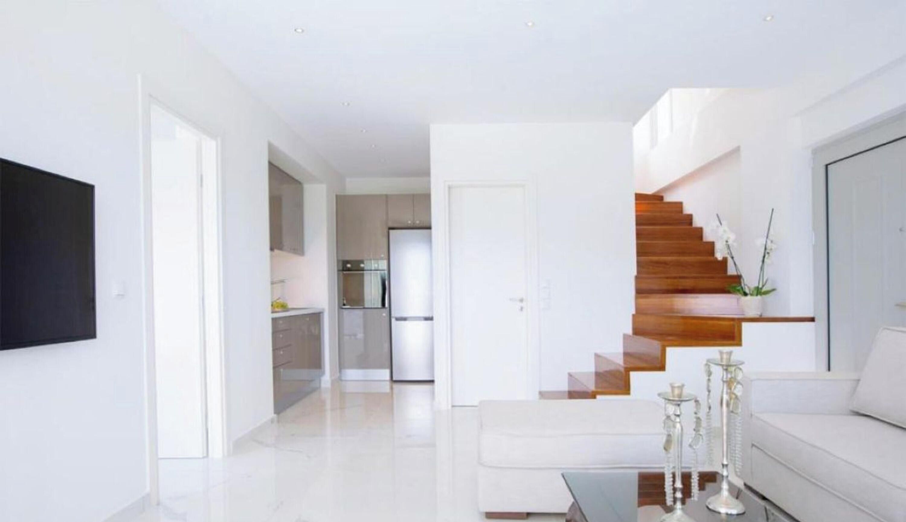 A modern living room with a staircase, a kitchen in the background, and a flat-screen TV mounted on the wall.