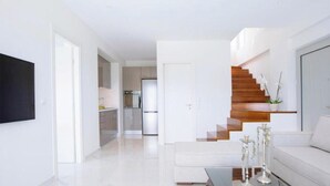 A modern living room with a staircase, a kitchen in the background, and a flat-screen TV mounted on the wall. - Omicron & White Villas (Lefkada)