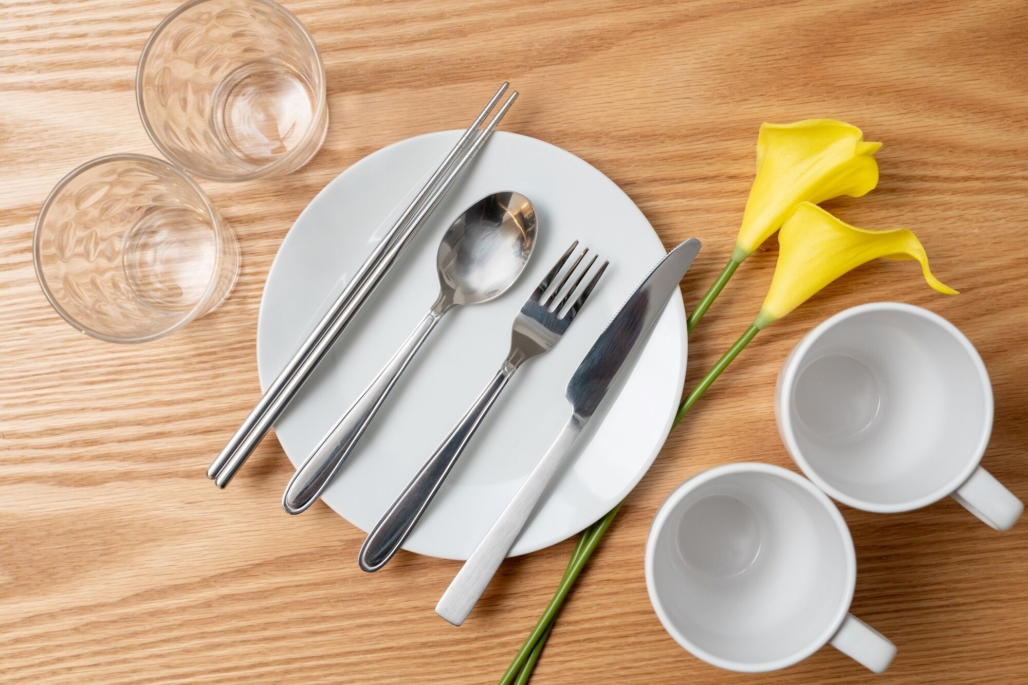 A neatly arranged dining setup with a plate, cutlery, two glasses, and a yellow flower on a wooden table.