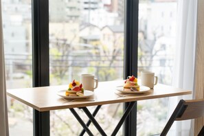 A table with two plates of dessert and two cups, positioned in front of a window with a view of buildings.