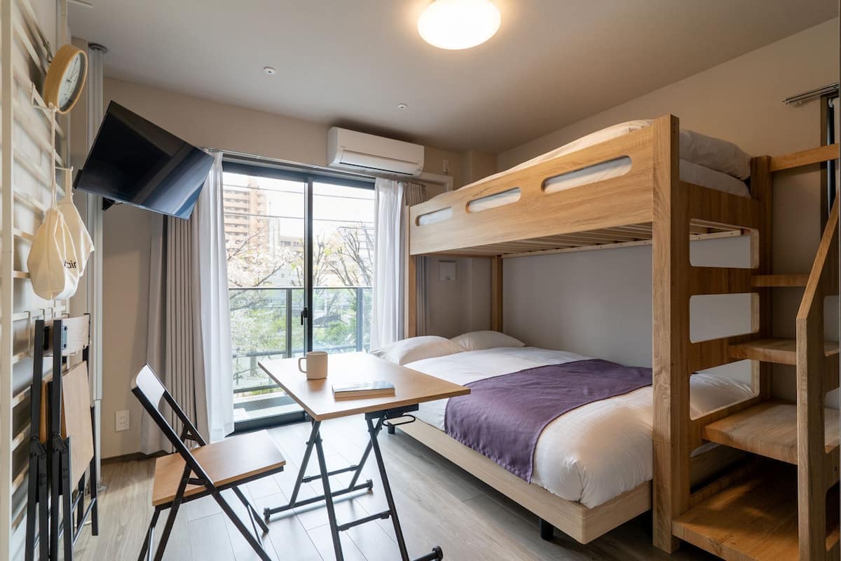 A bunk bed room with a desk and chair, a window with a view of buildings, and an air conditioning unit.
