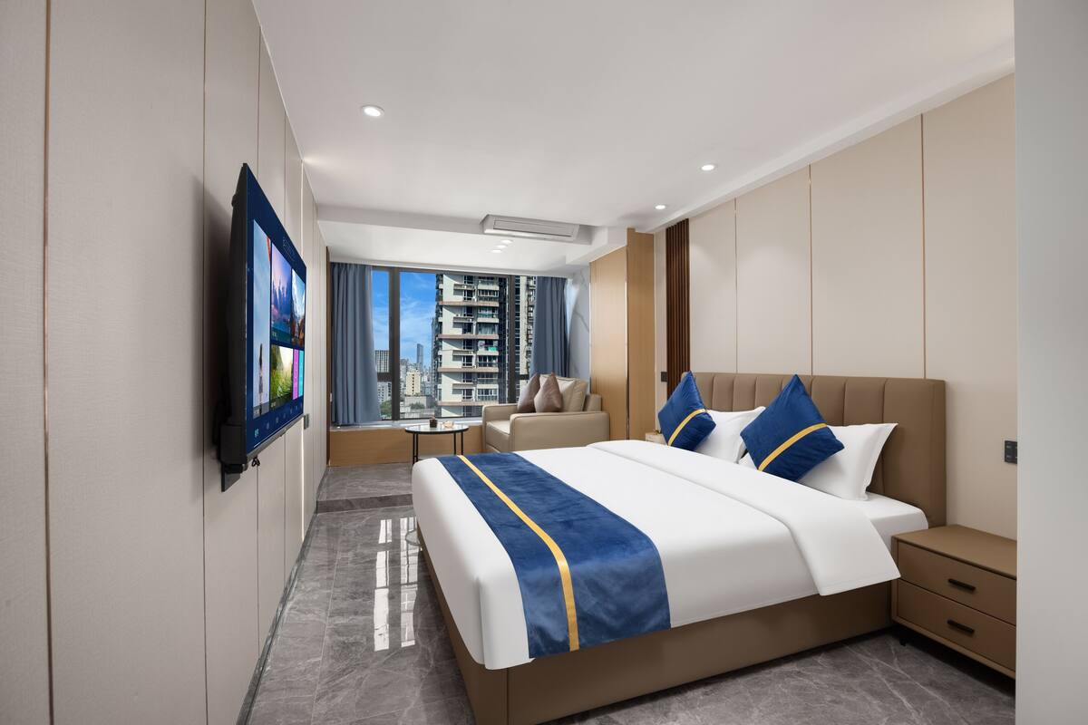 A modern hotel room with a large bed, a TV mounted on the wall, a seating area with a table and chairs, and a view of the cityscape through large windows.