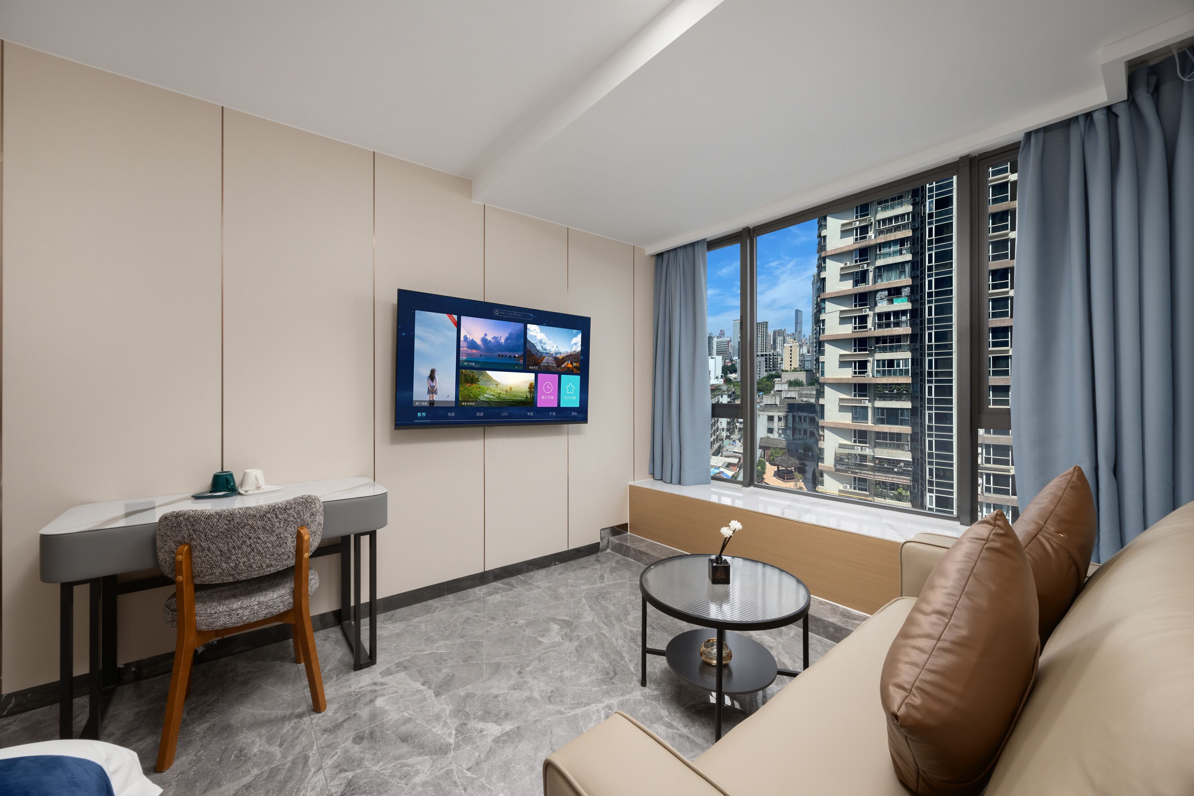 A modern hotel room with a flat-screen TV mounted on the wall, a dining area with a table and chairs, a round coffee table, and a sofa.