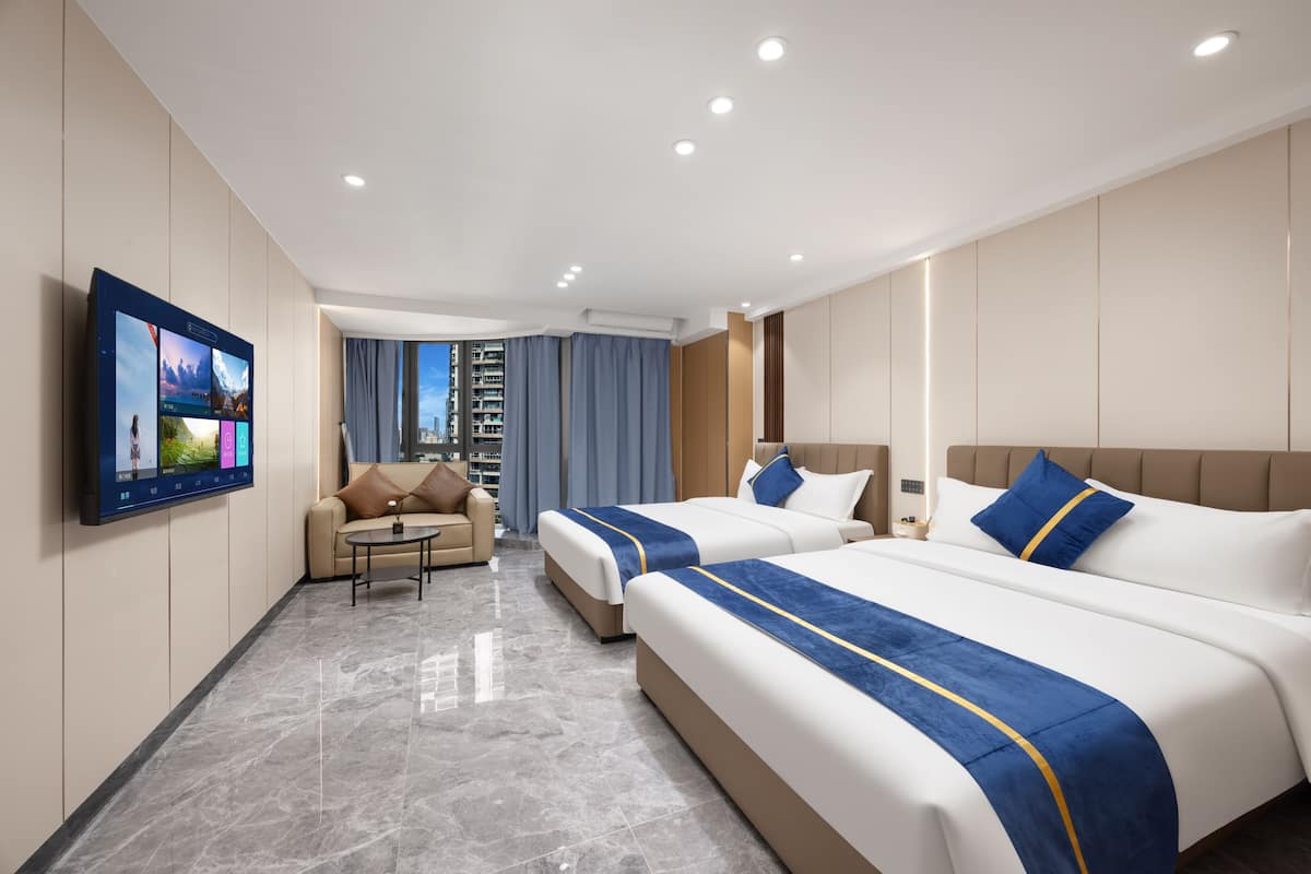 A hotel room with a large flat-screen TV, two beds with blue and yellow bedspreads, a sofa, a small table, and a view of a cityscape.