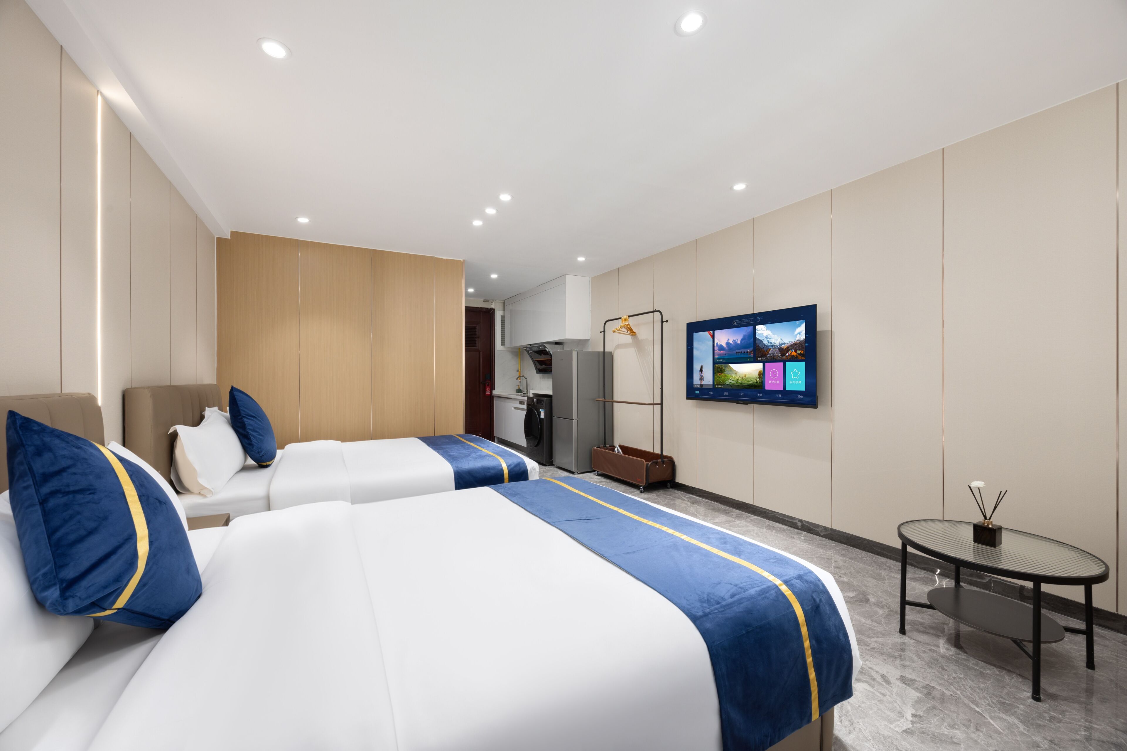 A hotel room with two beds, a television on the wall, a small table, and a kitchenette.