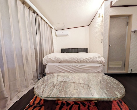 A hotel room with a bed, a glass-top table, a door, and a window with curtains.