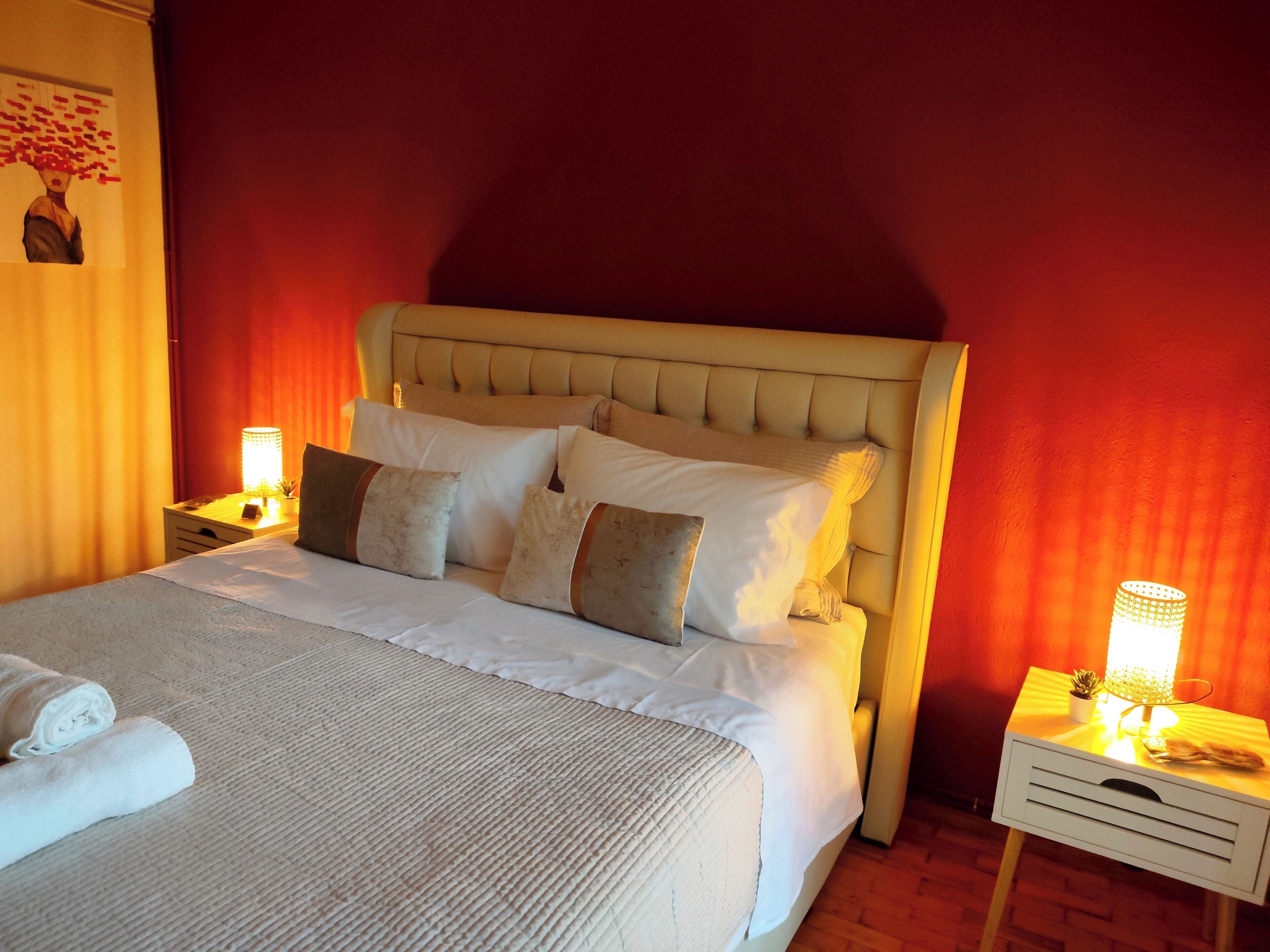 Un lit bien fait, avec une couette blanche et plusieurs oreillers, une table de chevet avec une lampe, et un mur sur lequel est accroché un rideau rouge.