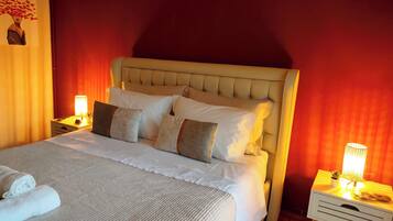 Un lit bien fait, avec une couette blanche et plusieurs oreillers, une table de chevet avec une lampe, et un mur sur lequel est accroché un rideau rouge.