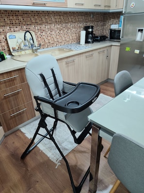 A kitchen with a high chair, a refrigerator, a coffee maker, and a tiled backsplash.