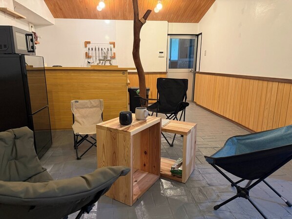 Ein Zimmer mit Holzboden, einem Schreibtisch mit Stuhl, einem schwarzen Klappstuhl, einem kleinen Tisch mit Tasse und einem Kühlschrank.