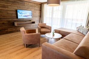 Ein modernes Wohnzimmer mit Holzwänden, einem Flachbildfernseher, einer Couch, zwei Sesseln, einem kleinen Tisch und einem großen Fenster mit Vorhängen.
