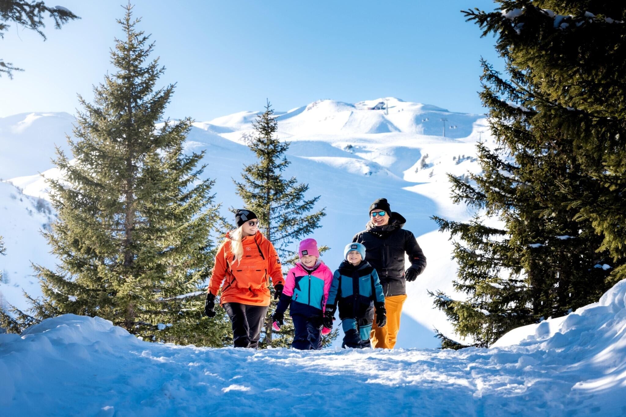 A group of people walking on a snowy path with snow-covered mountains and evergreen trees in the background.