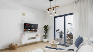 Een moderne woonkamer met een flatscreen televisie, een witte salontafel, een potplant en een schuifdeur die leidt naar een buitenruimte met een bankje en planten.
