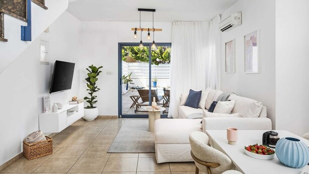 Een moderne woonkamer met een bank, salontafel en televisie. Links is een trap te zien en er is uitzicht op een terras buiten met planten en meubels.