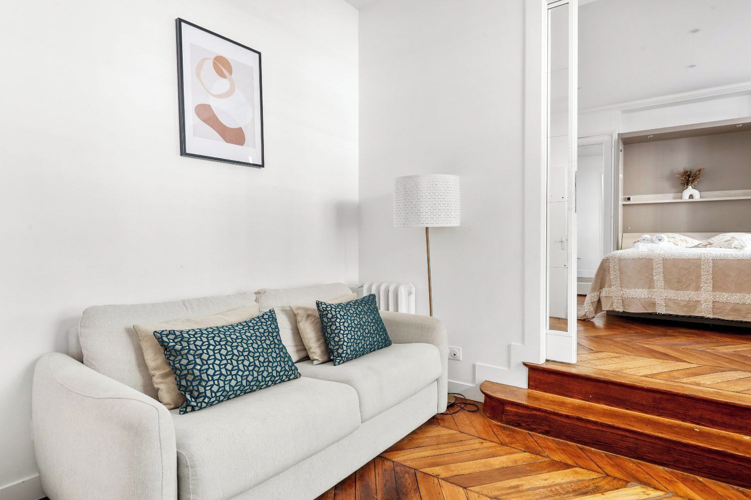 A modern living room with a sofa, patterned cushions, a framed artwork, a floor lamp, and a view into a bedroom with a bed and a shelf.