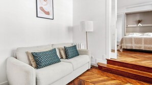 A modern living room with a sofa, patterned cushions, a framed artwork, a floor lamp, and a view into a bedroom with a bed and a shelf. - Bright Parisian 1BR by Champs (Paris)