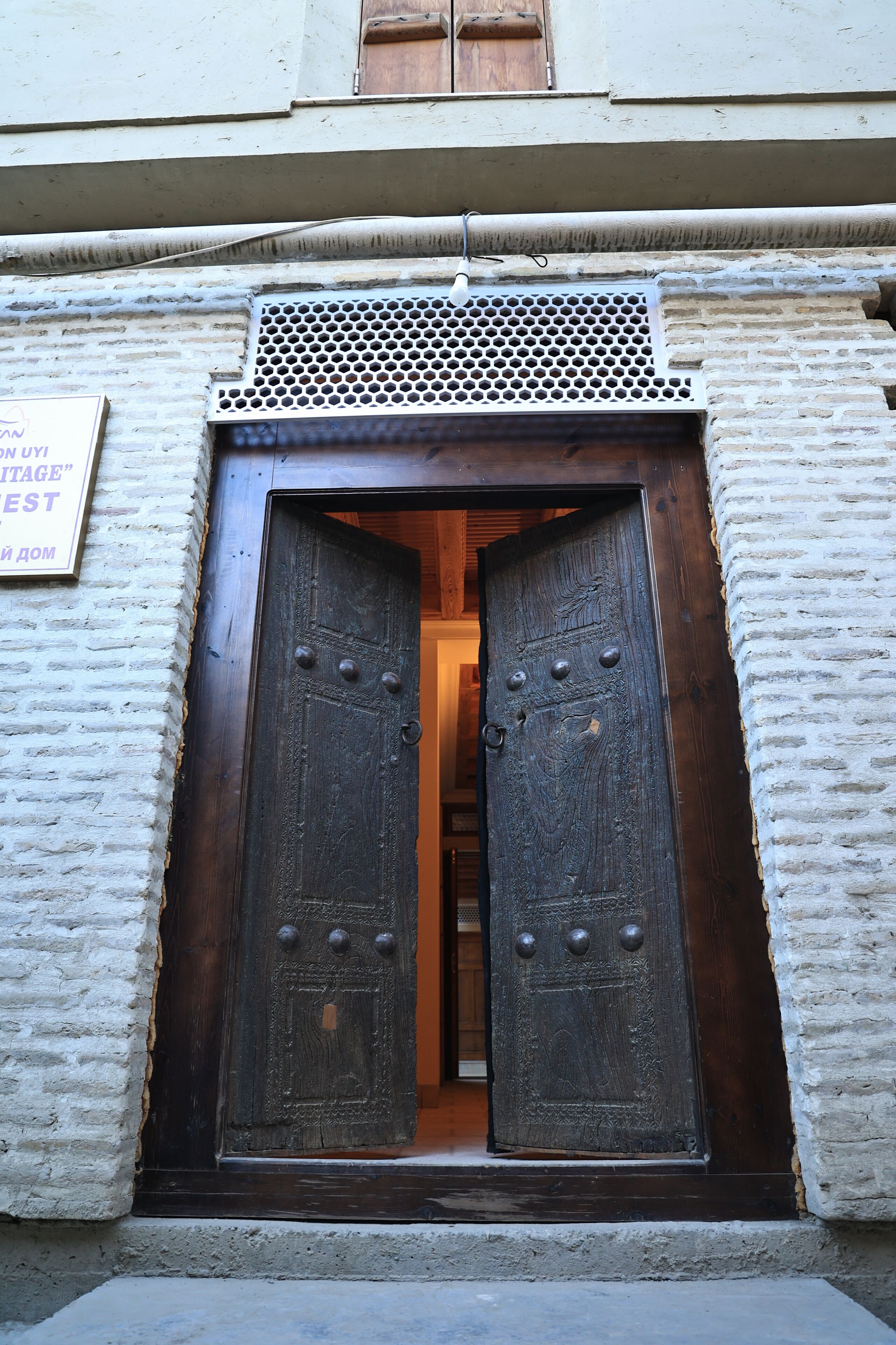 Una puerta de madera grande con una celosía de metal en la parte superior, ubicada en un edificio de paredes de ladrillo blanco.