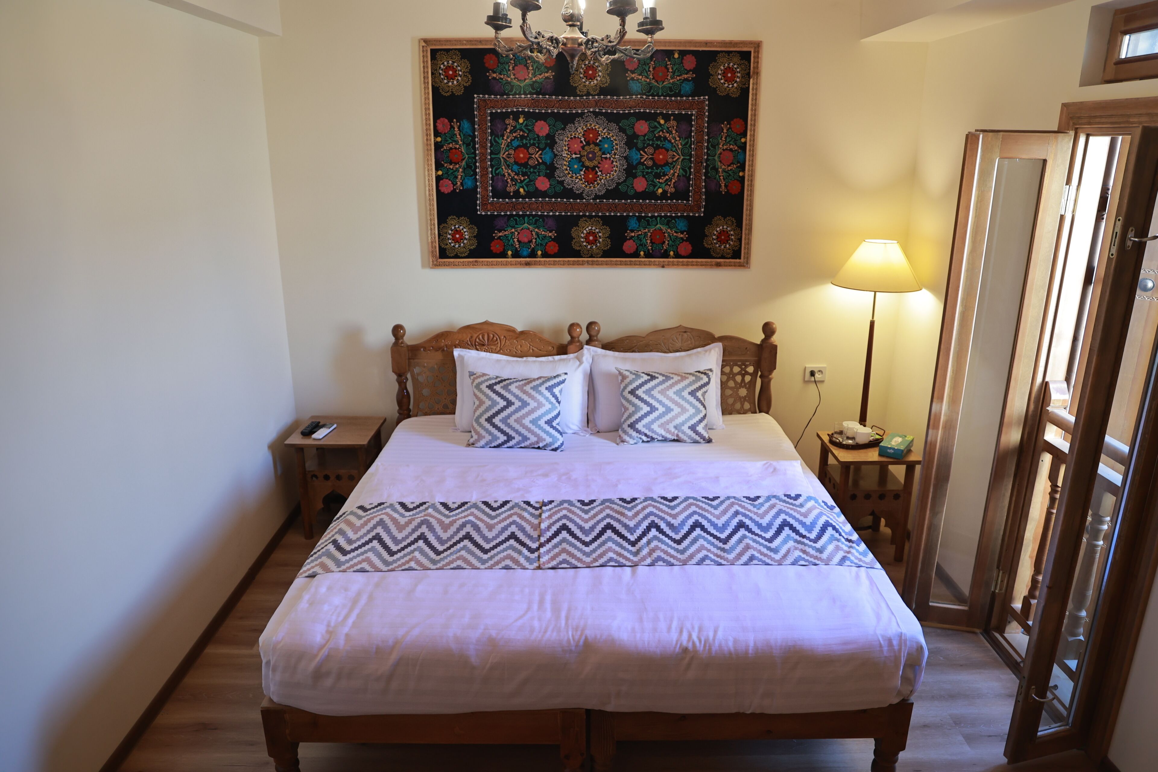 A bed with a patterned bedspread, a wall hanging, a bedside lamp, and a wooden nightstand with a phone.