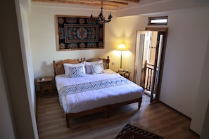 Quarto com cama de madeira, mesa de cabeceira, luminária, tapeçaria decorativa e porta que dá para outro cômodo.