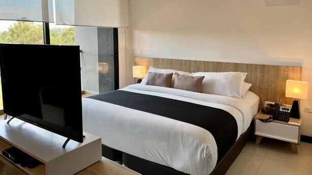 A modern hotel room with a large bed, a flat-screen TV, bedside lamps, and a window with a view. - Rioverde Living suites- Apartahotel (Antioquia)