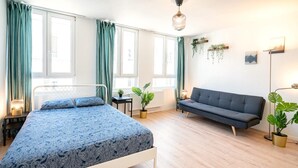 A bedroom with a bed, sofa, two windows, a radiator, and potted plants.