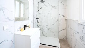 A bathroom with a white marble shower, a white sink with a mirror, and a beige floor.