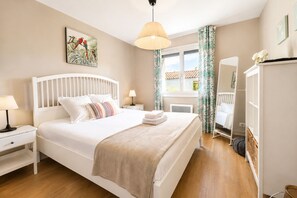 Quarto com cama, mesas de cabeceira, luminária pendente, janela com cortinas, radiador, guarda-roupa e uma obra de arte na parede.