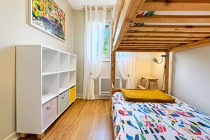 Ein Zimmer mit einem Etagenbett, einem weißen Kleiderschrank, einer farbenfrohen Bettdecke, einem Fenster mit Vorhängen und einem Bild an der Wand.