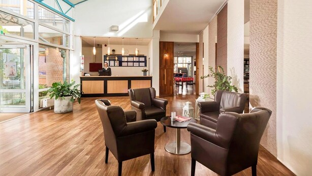 Eine Hotellobby mit Ledersesseln, einem Empfangstresen und einem Eingang mit Glastür.