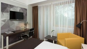 Ein Hotelzimmer mit Bett, Schreibtisch mit Fernseher, Sessel, kleinem Tisch mit Lampe und einer Couch.