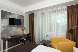 A hotel room with a bed, a desk with a television, a chair, a small table with a lamp, and a sofa.