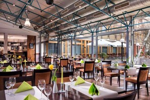 Un restaurante con techo alto, vigas a la vista y amplias ventanas. Las mesas están dispuestas con servilletas verdes, copas de vino y velas.