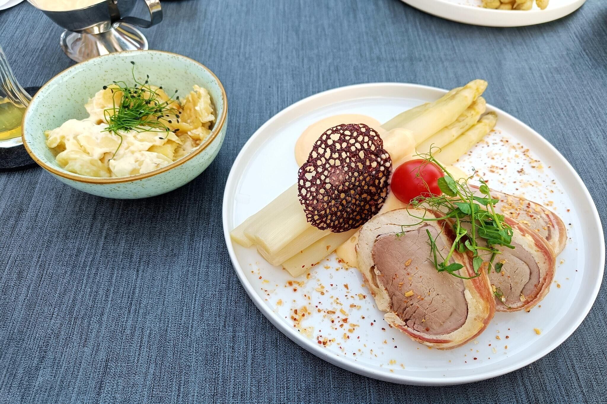 A plate with sliced meat, a round bread roll, a cherry tomato, and a bowl of potato salad.