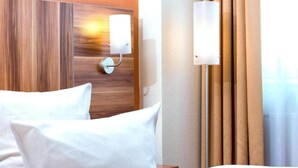 Ett hotellrum med en sänggavel i trä, en säng med vita sängkläder, en väggmonterad lampa och ett fönster med gardiner.
