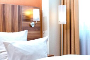 Ein Hotelzimmer mit einem hölzernen Kopfende, einem Bett mit weißen Leinen, einer Wandleuchte und einem Fenster mit Vorhängen.