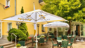 Un patio al aire libre con sillas y mesas de mimbre verde, sombrillas blancas y fachada de un edificio amarillo.