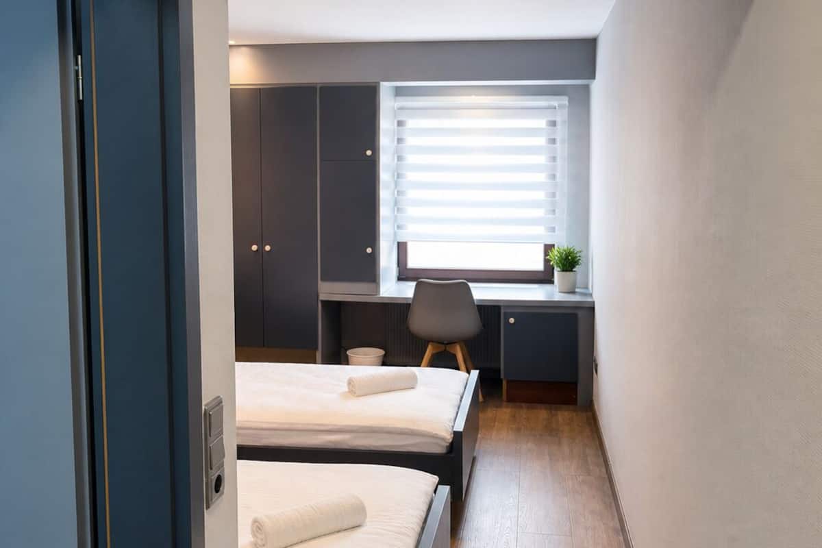 A compact room with two beds, a desk, and a window with blinds.