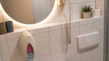 A bathroom with a round mirror, a hand soap dispenser, a toilet paper holder, and a small potted plant.