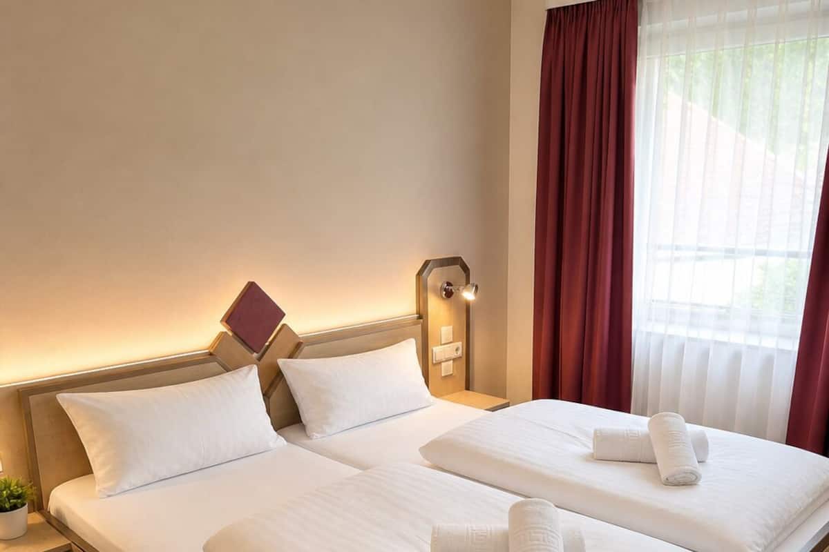 A double bed with white linens, a bedside table with a lamp, and a window with curtains.