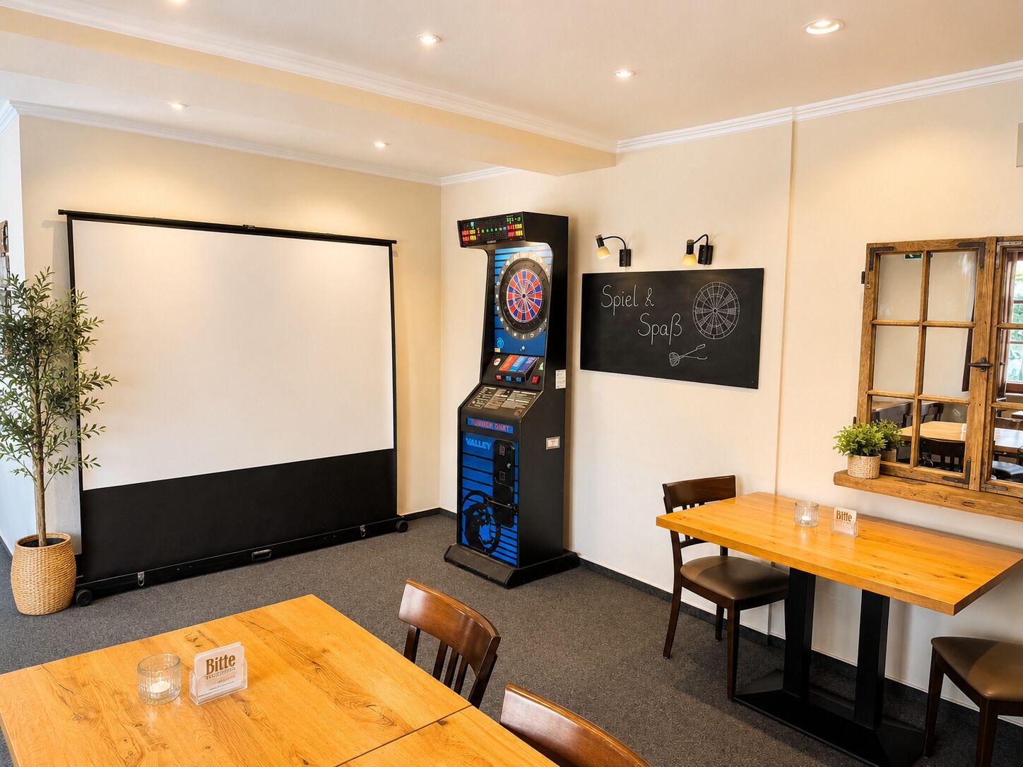 A room with a dartboard, a billiards machine, a chalkboard with writing, a wooden table, and chairs.