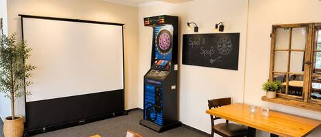 A room with a dartboard, a billiards machine, a chalkboard with writing, a wooden table, and chairs.