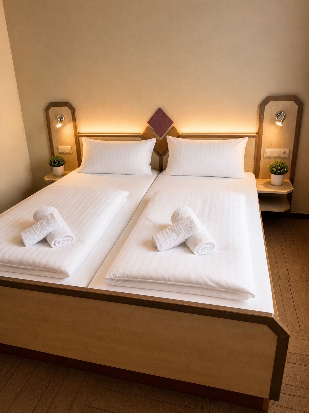 Cama doble con ropa de cama y toallas blancas, cabecera de madera, mesitas de noche con lámparas y una planta pequeña a cada lado.