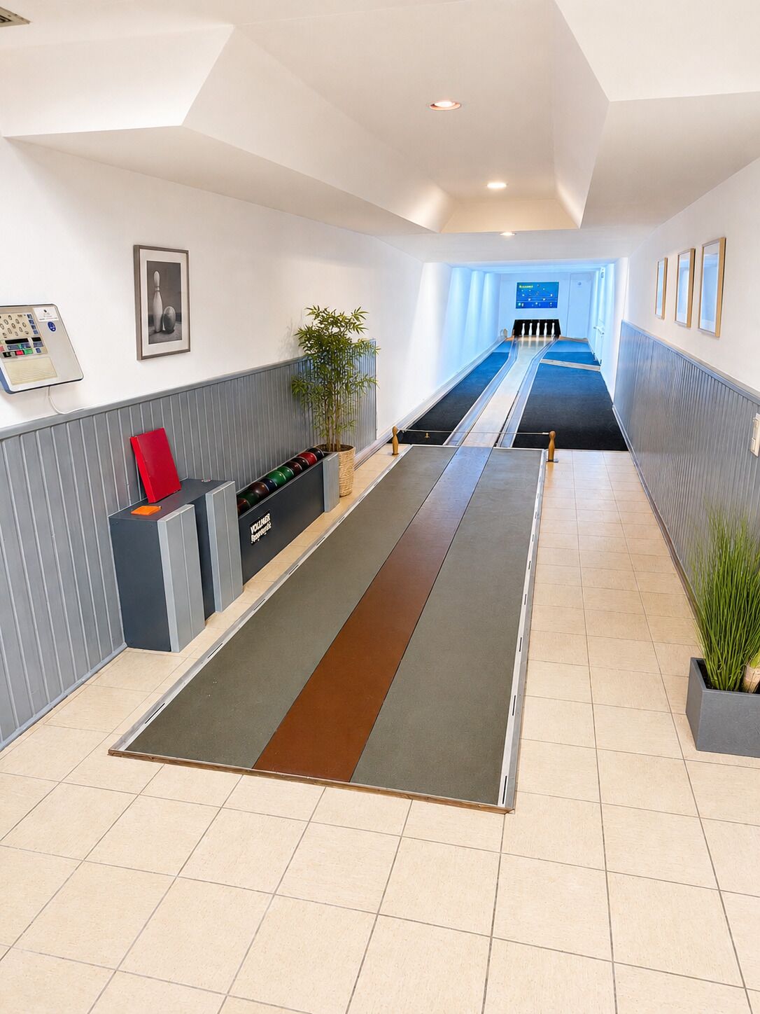 Een moderne hotelentree met een receptiebalie, een digitale informatiezuil en een lange gang die naar een zwembadgedeelte leidt.